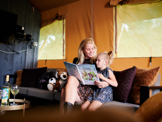 Een moeder en dochter lezen samen gezellig een boek op de comfortabele bank in het sfeervolle interieur van een safaritent.