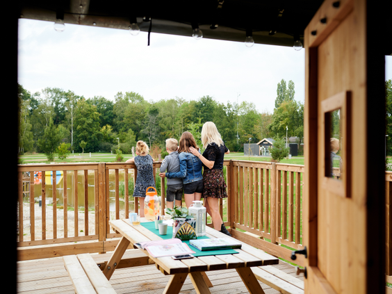 Uitzicht vanuit de safaritent op een gezin dat geniet van een drankje aan de picknicktafel op hun ruime, houten veranda aan het water.