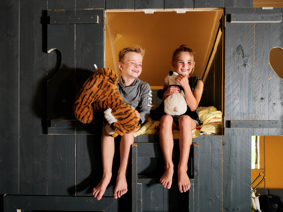 Blije broer en zus zitten met hun knuffels in de opening van de gezellige, donkerhouten bedstee in een safaritent op Sallandshoeve.