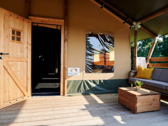 De openstaande houten deur van een safaritent geeft zicht op de overdekte veranda met een comfortabele loungebank.