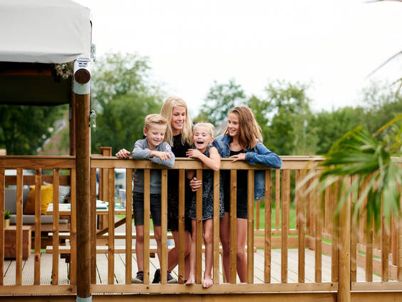 Een lachend gezin met drie kinderen leunt over de houten balustrade van de veranda van hun safaritent op Vakantiepark Sallandshoeve.