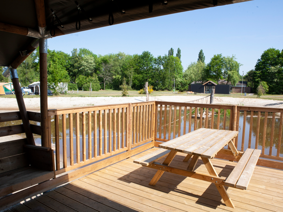 Zonnig uitzicht vanaf de houten veranda met picknicktafel op de speelvijver en de groene omgeving van Vakantiepark Sallandshoeve.