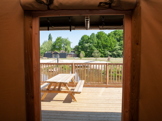 Het zonnige uitzicht vanuit een safaritent op de houten veranda met picknicktafel, direct gelegen aan de speelvijver van Vakantiepark Sallandshoeve.