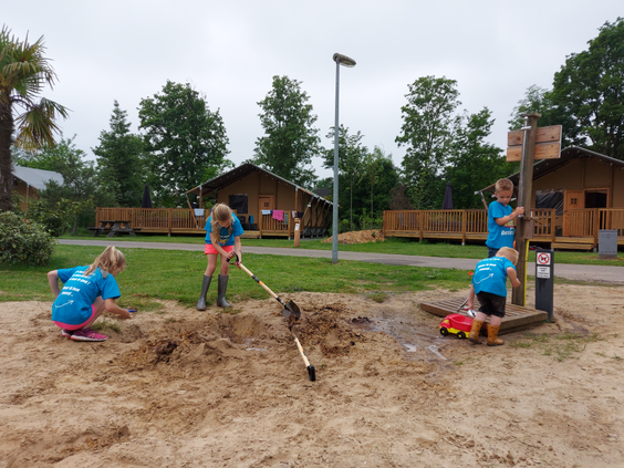 Drie kinderen in blauwe shirts spelen en graven enthousiast met scheppen en een speelgoedauto in de grote zandspeeltuin met waterpomp op Vakantiepark Sallandshoeve.
