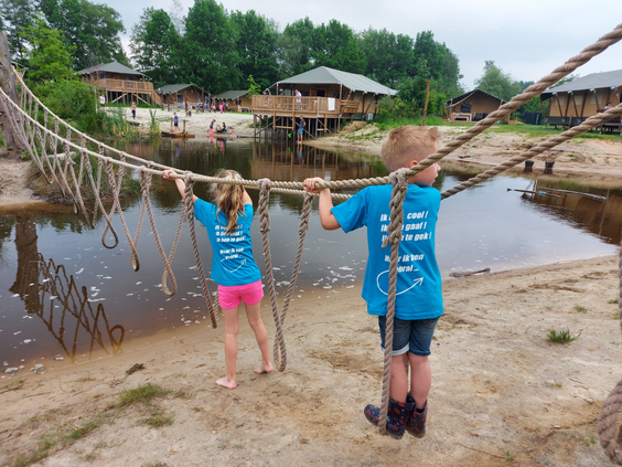 Twee kinderen in blauwe shirts balanceren voorzichtig over de wiebelende touwbrug die over de speelvijver van Sallandshoeve loopt.