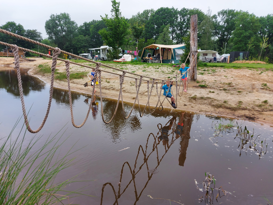 Overzicht van de natuurlijke speelvijver op Vakantiepark Sallandshoeve, met kinderen die over de uitdagende touwbrug en het vlot klimmen.