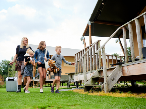 Een blij gezin met koffers en knuffels loopt enthousiast over het gras naar de trap van hun safarilodge op Sallandshoeve.