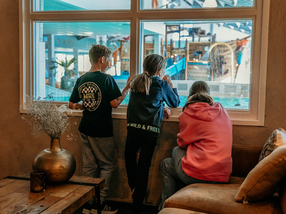 Drie kinderen kijken vanuit het restaurant door een groot raam naar de binnenspeeltuin van Strandpark De Zeeuwse Kust.