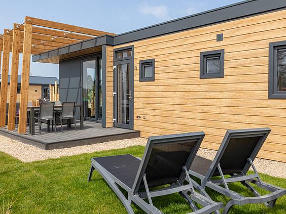 Buitenaanzicht van een modern houten chalet met een overdekt terras en twee ligbedden in de tuin op Strandpark De Zeeuwse Kust.