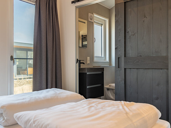 Ouderslaapkamer met een comfortabel opgemaakt bed en een doorkijk naar de moderne ensuite badkamer in een chalet op De Zeeuwse Kust.