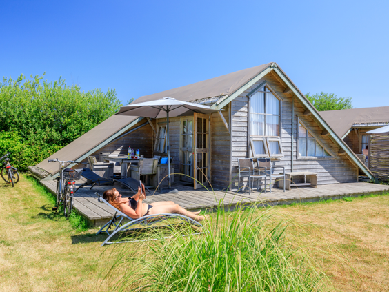 Een houten chalet met een ruim terras op een grasveld waar iemand ligt te zonnen op Strandpark De Zeeuwse Kust.