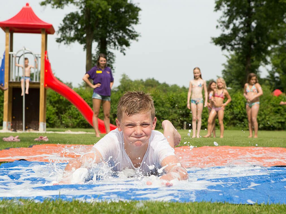 Een jongen glijdt met veel plezier op zijn buik over de lange buikschuifbaan op het gras van Camping 't Veld.