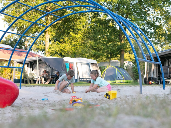 Twee kinderen spelen in de zandbak onder een blauw klimrek op de camping, vlak bij de kampeerplaatsen.