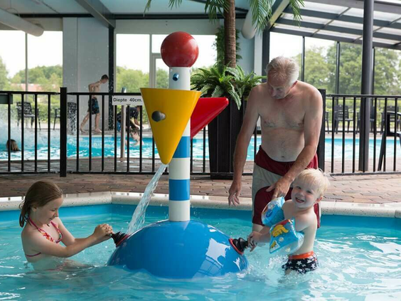 Een vader speelt met zijn jonge kinderen in het kinderbad bij een waterspuitend speeltoestel op Camping 't Veld.