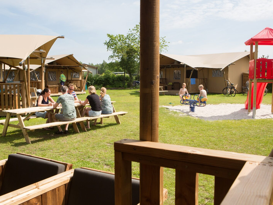 Gezinnen zitten gezellig buiten bij hun safaritenten op het glampingveld, terwijl kinderen spelen in de centrale speeltuin.