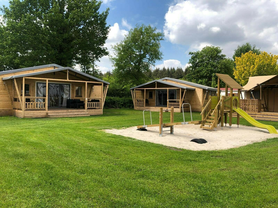 Moderne houten lodges op Camping 't Veld, gelegen aan een groot grasveld met een centrale speeltuin in de zandbak.