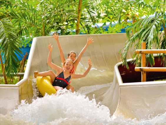 Twee blije meiden roetsjen met hun armen in de lucht in een gele band van een brede waterglijbaan in het zwemparadijs van Tropical Islands.