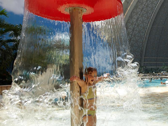 Een klein kind staat lachend onder een grote waterpaddenstoel die als een waterval van water naar beneden stort in het kinderbad van Tropical Islands.
