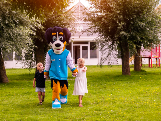 De mascotte van Vakantiepark Aquadelta wandelt hand in hand met twee blije kinderen over een grasveld op het park.