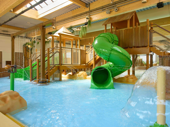 Overdekt zwemparadijs met een grote groene waterglijbaan en diverse houten speeltoestellen.