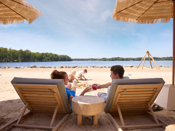 Koppel ontspant op strandstoelen onder een parasol aan het Victoriameer met twee kinderen dat in het zand aan het spelen zijn