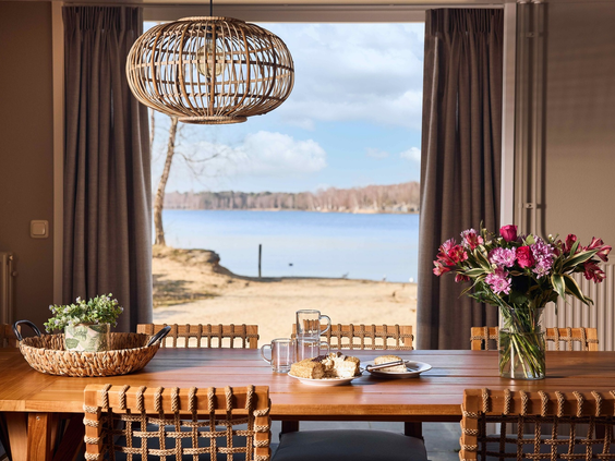 Eetkamer met een houten tafel gedekt voor een maaltijd en een groot raam dat uitkijkt op het strand en het meer