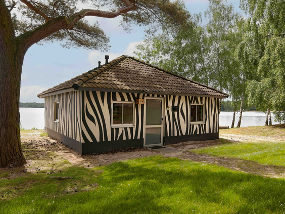 Buitenaanzicht van de vrijstaande 5 persoons Forest Jungalow met zebrapatroon in een bosrijke omgeving aan het water in Lake Resort Beekse Bergen