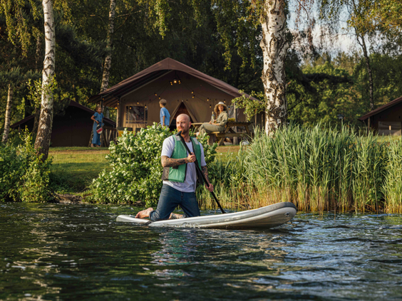 Man met een zwemvest is aan het suppen op het meer met op de achtergrond een safaritent en bomen in Lake Rsort Beekse Bergen