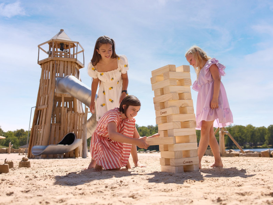 Kinderen spelen met reuze Jenga op het zandstrand van Vakantiepark Beekse Bergen, met een houten speeltoren op de achtergrond.