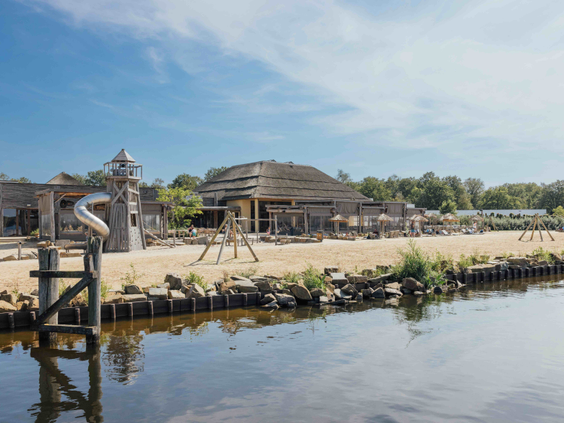 Uitzicht vanaf het water op het speelstrand en centrumgebouw Ganvie Village van Lake Resort Beekse Bergen