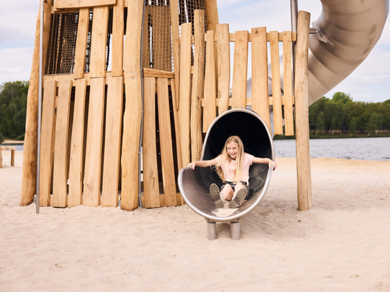Een meisje zit lachend in de ronde opening van een metalen tunnelglijbaan in een houten speeltuin op de Beekse Bergen.