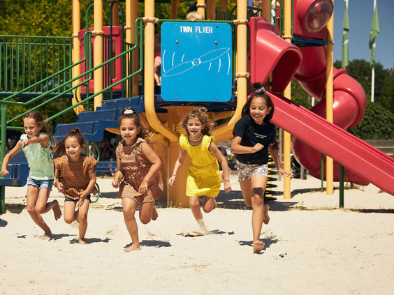 Vijf jonge kinderen rennen en lachen in een zandspeeltuin bij Lake Resort Beekse Bergen.