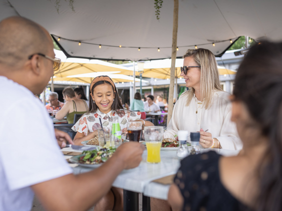 Een lachende familie geniet samen van een lunch met drankjes op het zonnige terras van het restaurant op Bospark 't Wolfsven.