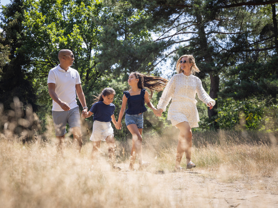 Een gezin met twee dochters rent en wandelt vrolijk hand in hand door de bosrijke, zonnige natuur van Bospark 't Wolfsven.