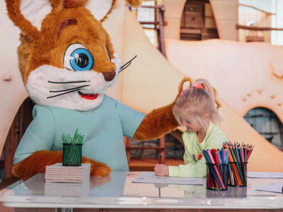 Een meisje is aan het kleuren en knutselen aan tafel met de vrolijke parkmascotte Koos in de kidsclub van Bospark 't Wolfsven.