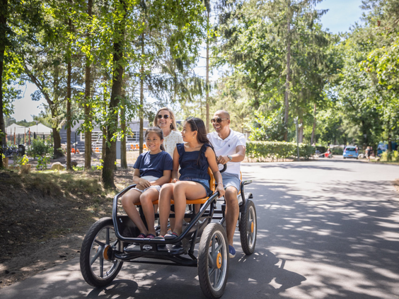 Een gezin van vier personen rijdt vrolijk op een familiefiets (skelter) door de bosrijke omgeving van Vakantiepark Bospark 't Wolfsven.