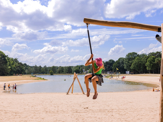 Een vrolijk meisje schommelt boven het zandstrand, met het recreatiemeer van Bospark 't Wolfsven op de achtergrond.
