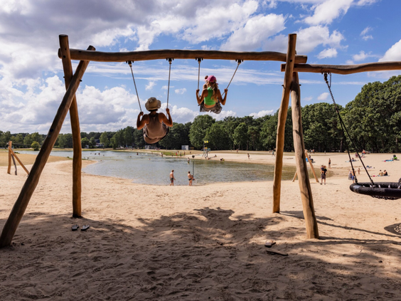 Twee kinderen schommelen naast elkaar op de houten schommels op het zandstrand van Bospark 't Wolfsven in Noord-Brabant.