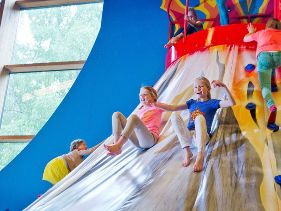 Kinderen klimmen en glijden van een grote, zilverkleurige glijbaan in de binnenspeeltuin.