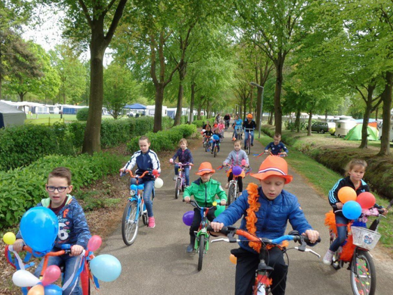 Grote groep kinderen doet mee aan de optocht met versierde fietsen, georganiseerd door het animatieteam.