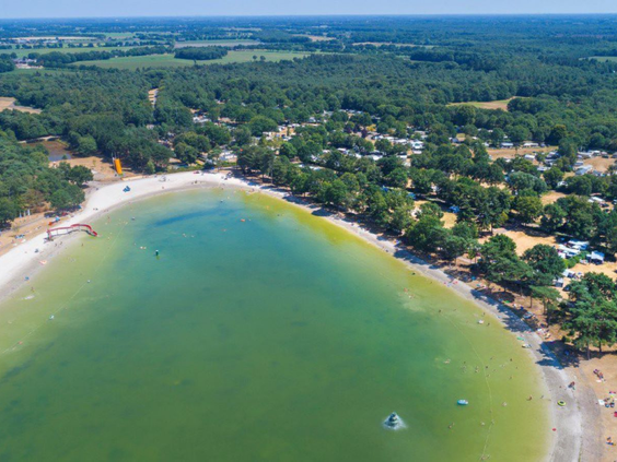 Helderblauw water en een uitgestrekt zandstrand bij de recreatieplas van Vakantiepark De Bergen, gezien vanuit de lucht.