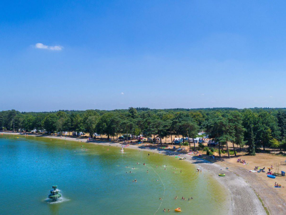 Weids overzicht van de grote recreatieplas met zandstrand, waterglijbaan en speeltoestellen in het water.