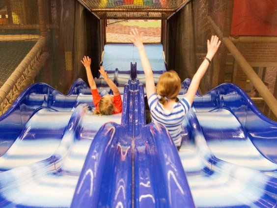 Twee kinderen met de handen in de lucht glijden van een snelle blauwe glijbaan in de binnenspeeltuin van Vakantiepark De Bergen.