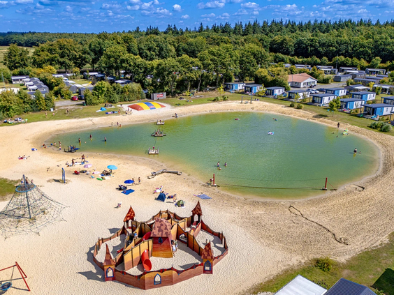 Overzicht van de zwemvijver met zandstrand en speeltoestellen op de camping