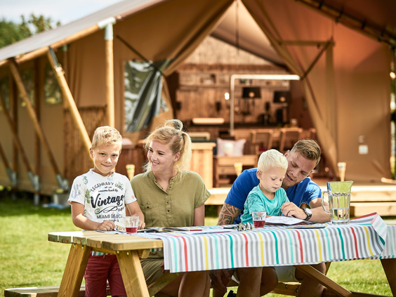 Een gezin met twee kinderen zit gezellig aan een picknicktafel voor hun luxe safaritent op de glamping van Vakantiepark De Krim.