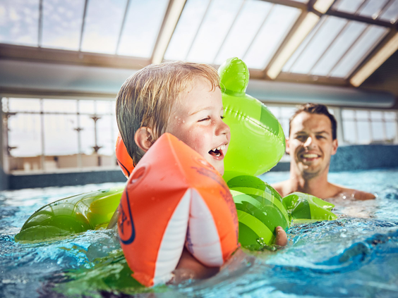 Een lachende peuter met zwemvleugels speelt samen met zijn vader in het binnenzwembad van Vakantiepark De Krim.