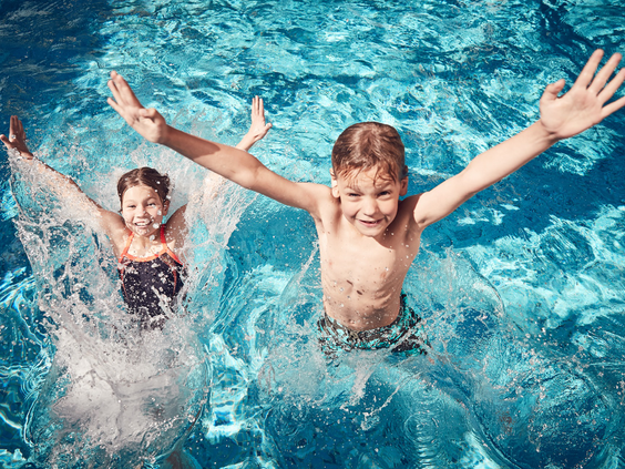 Twee kinderen, een jongen en een meisje, springen met een grote plons in het helderblauwe water van het buitenzwembad.