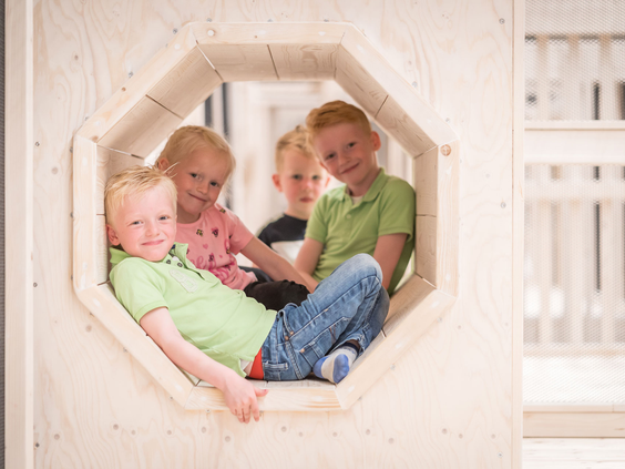 Vier kinderen kijken en lachen vanuit een achthoekig raam in een houten speeltoestel in de binnenspeeltuin.