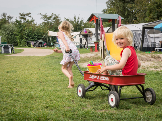 Een meisje trekt een lachend jongetje in een rode bolderkar over het grasveld van de camping bij Vakantiepark De Krim.