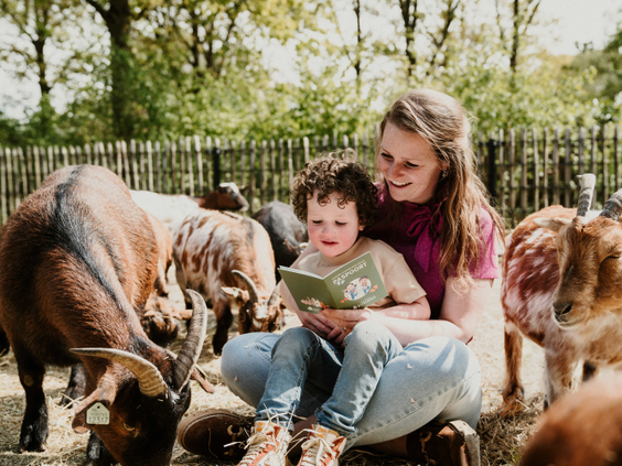 Een moeder leest haar zoontje voor uit een boek, terwijl ze op de grond zitten omringd door geitjes op de kinderboerderij van Vakantiepark Dierenbos.